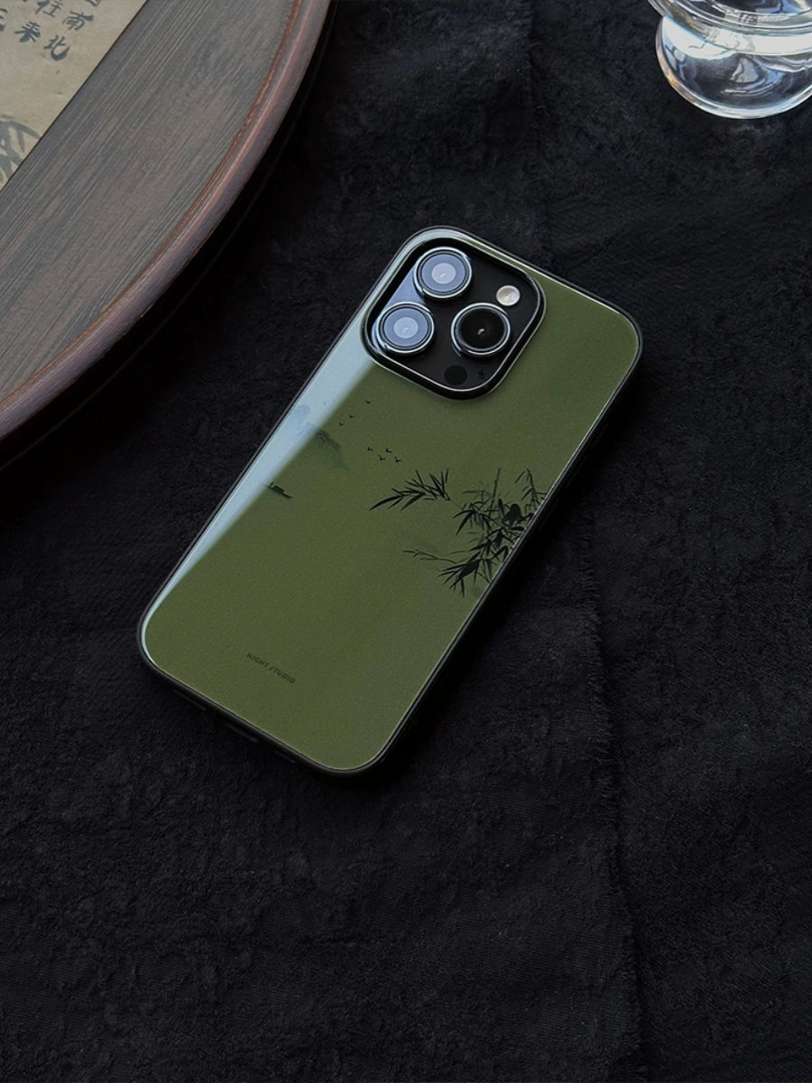 A green phone case with a river and mountain landscape painting, placed on a dark surface next to a glass with water and some plants.