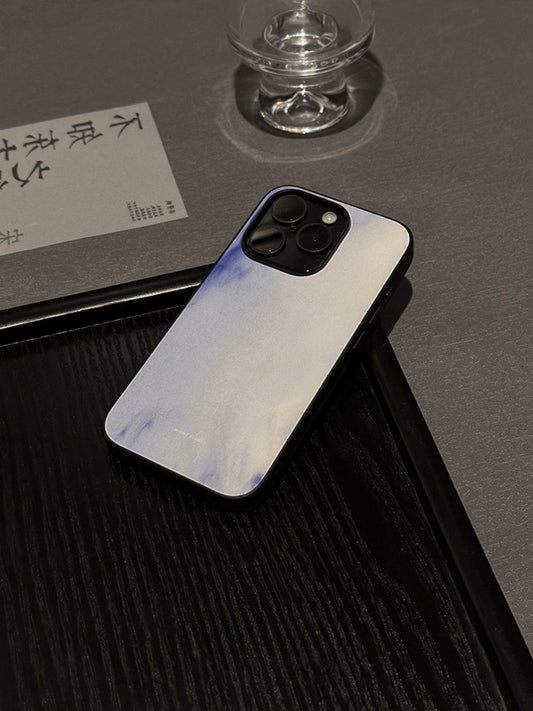 A phone case with a cloud design in shades of purple and white, placed on a black surface next to a glass.