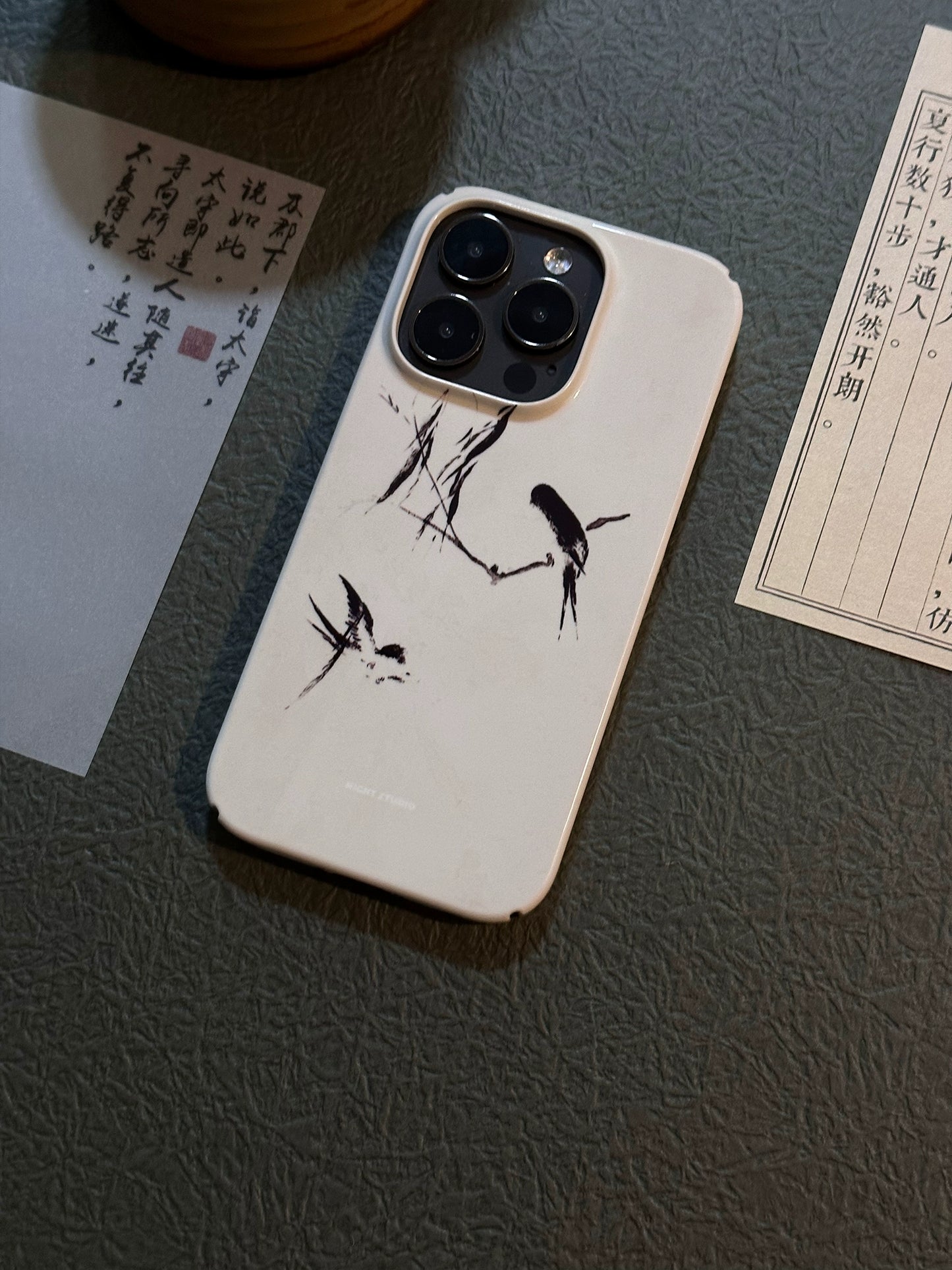 A white phone case with a black swallow pattern, displayed on a flat surface with documents and a pen in the background.