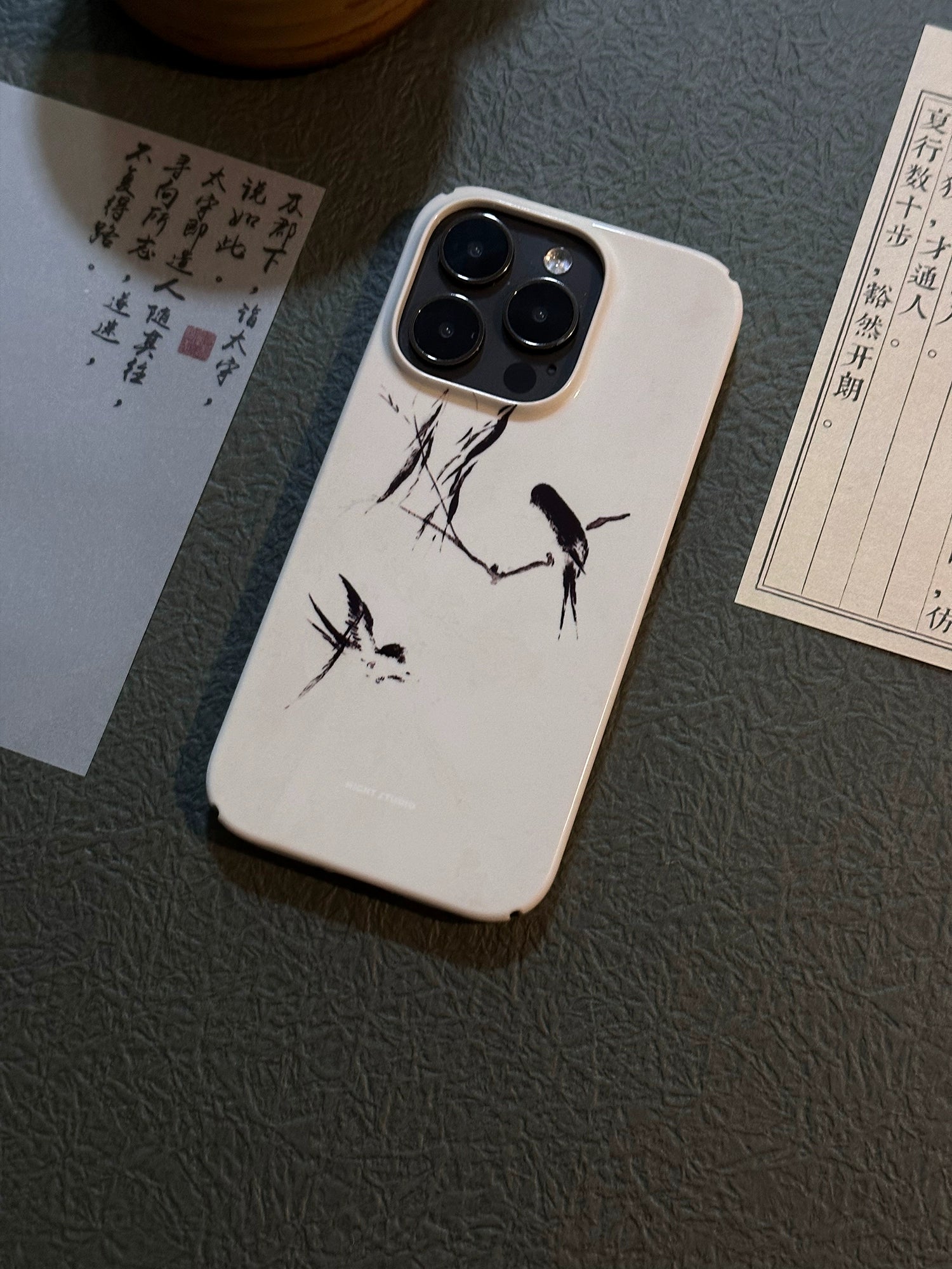A white phone case with a black swallow pattern, displayed on a flat surface with documents and a pen in the background.